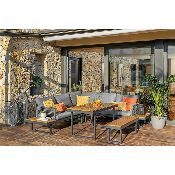 Conjunto de muebles Magalí - 5 pzs., Madera de acacia/Aluminio, Antracita - Acogedor conjunto de exterior con sofá gris, mesa de madera y bancos en una terraza con pared de piedra, perfecto para relajarse.