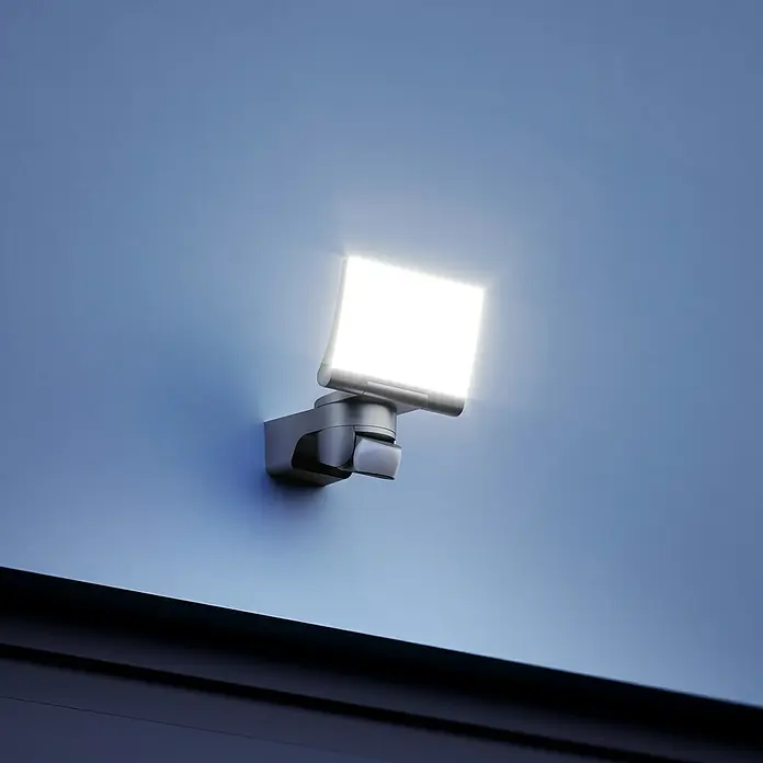 Steinel LED-Sensor-Strahler XLED home 2 S graphit - 13,7 W, L x B x H: 161 x 180 x 218 mm, Graphit, IP44 - Nahaufnahme eines LED-Strahlers mit Bewegungsmelder an einer blauen Wand, der ein helles, weißes Licht ausstrahlt.