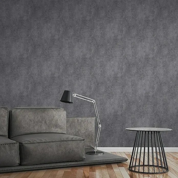 A.S. Création Titanium 3 Vliestapete I - Anthrazit, Betonoptik, 10,05 x 0,53 m - Graues modulares Sofa mit Steppdetails auf grauem Teppich, Akzentlampe und Beistelltisch vor einer dunklen Wand.