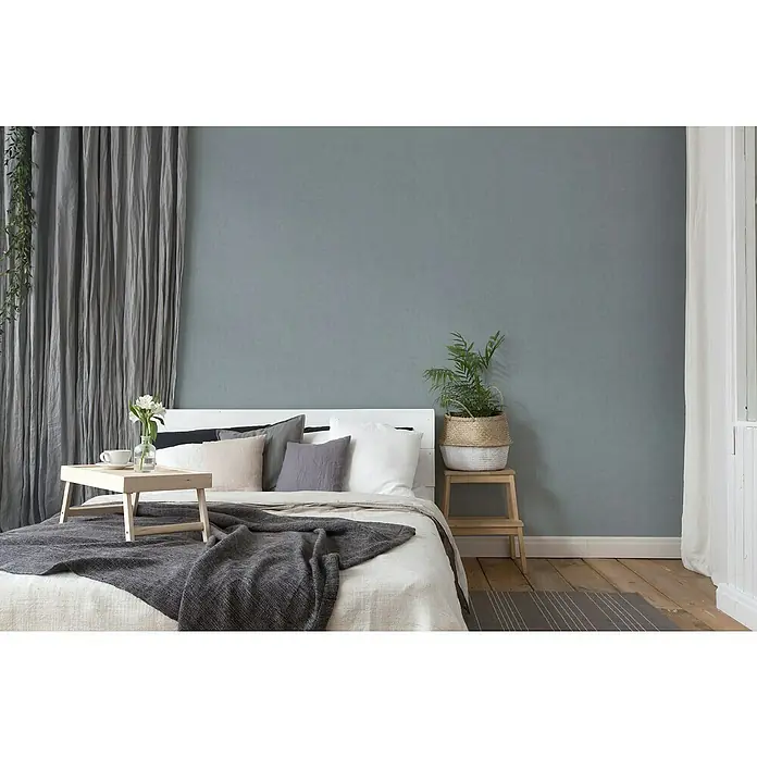 A.S. Création Titanium 3 Vliestapete - Pastellblau, Uni, 10,05 x 0,53 m - Schlafzimmer mit grauer Tagesdecke auf dem Bett. Ein Holztisch steht auf dem Bett und ein Hocker neben dem Bett mit Pflanze.