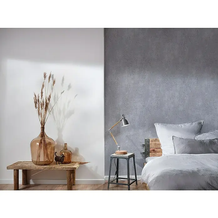 A.S. Création Titanium 3 Vliestapete I - Dunkelgrau, Betonoptik, 10,05 x 0,53 m - Einrichten mit einer rustikalen Holzbank, einer Glasvase mit Trockenblumen und einer Stehlampe auf einem Beistelltisch neben einem Bett.