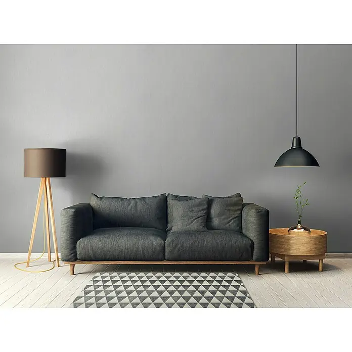 A.S. Création Daniel Hechter 6 Vliestapete Textilstruktur - Grau, Uni, 10,05 x 0,53 m - Eine stilvolle graue Couch mit Kissen, einer Stehlampe und einem Beistelltisch unter einer Hängelampe, die ein gemütliches Wohnzimmerambiente schaffen.