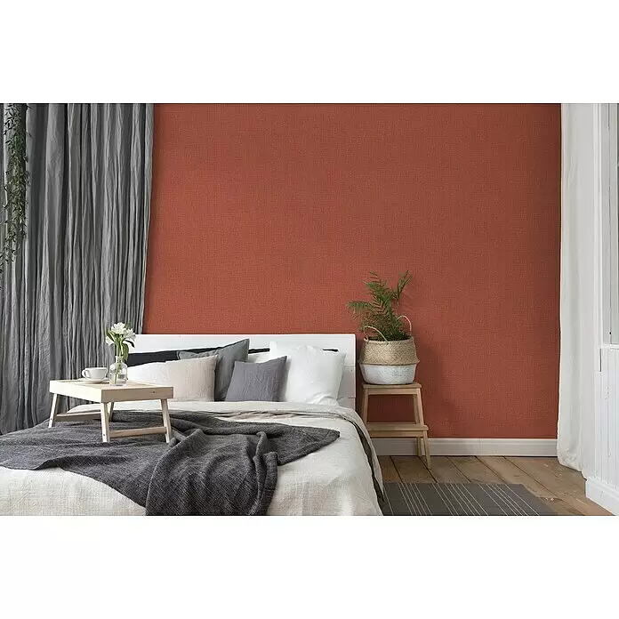 A.S. Création Desert Lodge Vliestapete - Rot, Uni, 10,05 x 0,53 m - Akzentwand aus roter strukturierter Tapete in einem gemütlichen Schlafzimmer mit Bett und Pflanzen.