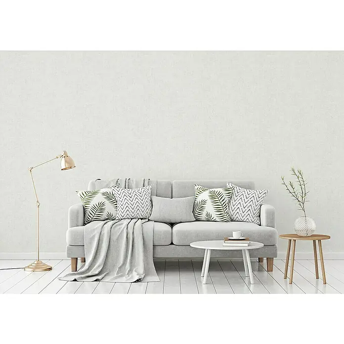 A.S. Création Titanium 3 Vliestapete - Strukturiertes Weiß, Uni, 10,05 x 0,53 m - Wohnzimmer mit modernem, grauem Sofa, Zierkissen mit Blattmotiven und geometrischen Mustern, einer Stehlampe und Beistelltischen.