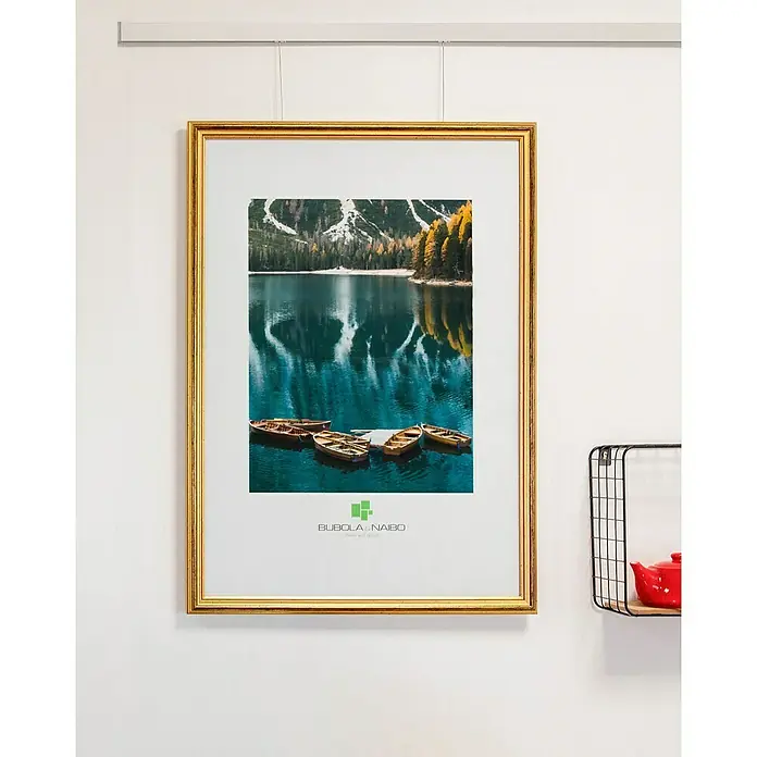 Bubola & Naibo Holzrahmen 2490 - 50 x 70 cm, Gold - Ein gerahmtes Bild mit einem goldenen Rahmen hängt an der Wand und zeigt einen See mit Booten und Bergen im Hintergrund.