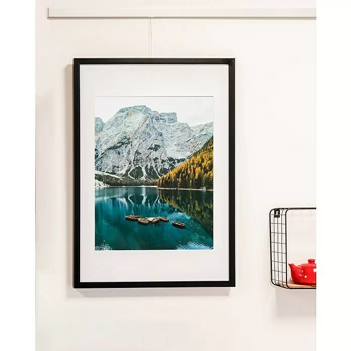 Nielsen Holzrahmen Apollo - 50 x 70 cm, Schwarz - Ein gerahmtes Foto eines Bergsees mit bunten Bäumen und Booten hängt an einer weißen Wand, neben einem schwarzen Drahtregal mit einer roten Teekanne.