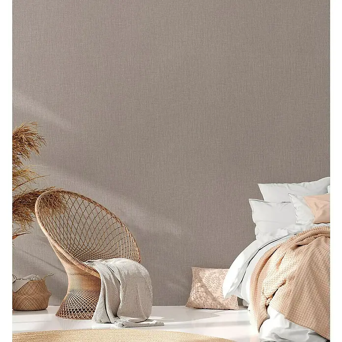A.S. Création Daniel Hechter 6 Vliestapete Textilstruktur - Beige, Uni, 10,05 x 0,53 m - Schlafzimmer-Interieur mit einem Rattan-Stuhl, einem stilvollen Bett und dekorativen Elementen, die eine gemütliche und entspannende Atmosphäre schaffen.