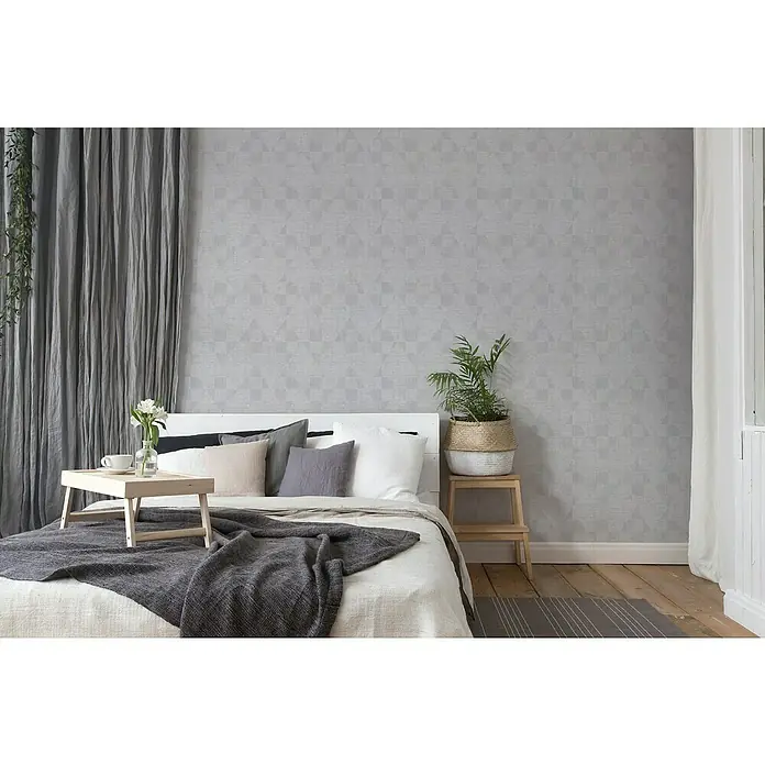 A.S. Création Titanium 3 Vliestapete - Silbergrau, Grafisch, 10,05 x 0,53 m - Ein Schlafzimmer mit einem Bett, das mit Kissen und einer grauen Decke dekoriert ist, sowie einem Holztischchen und einem Nachttisch mit einer Zimmerpflanze.