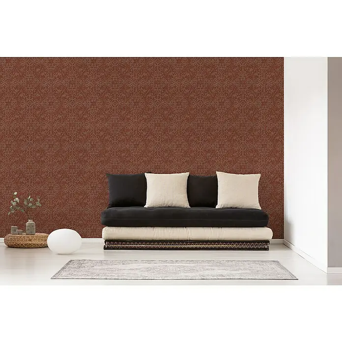 A.S. Création Desert Lodge Vliestapete Ethno-MusterRot, Grafisch, 10,05 x 0,53 m - Das Bild zeigt eine stilvolle Couch mit Kissen vor einer gemusterten Wand, hervorgehoben durch eine Lampe und einen Teppich.