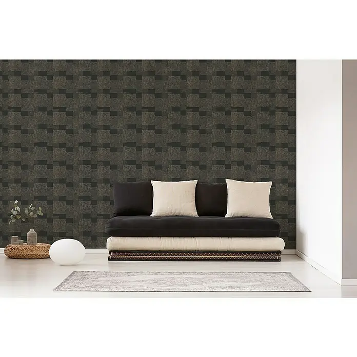 A.S. Création Desert Lodge Vliestapete Ethno-Muster - Schwarz/Gold, Grafisch, 10,05 x 0,53 m - Einrichtungsszene mit einem modernen Futonbett, schwarzen und beigen Kissen vor einer Wand mit abstraktem Muster. Die Beleuchtung ist weich.