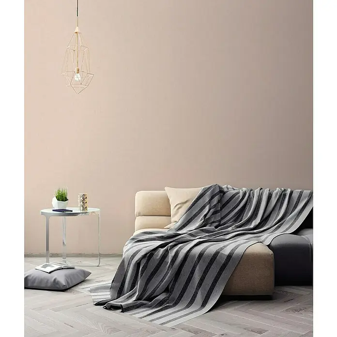 A.S. Création Titanium 3 Vliestapete - Wärmendes Beige, Uni, 10,05 x 0,53 m - Elegantes Wohnzimmer mit einem beigen Sofa, das mit einer gestreiften grauen Decke bedeckt ist und einer modernen Lampe.
