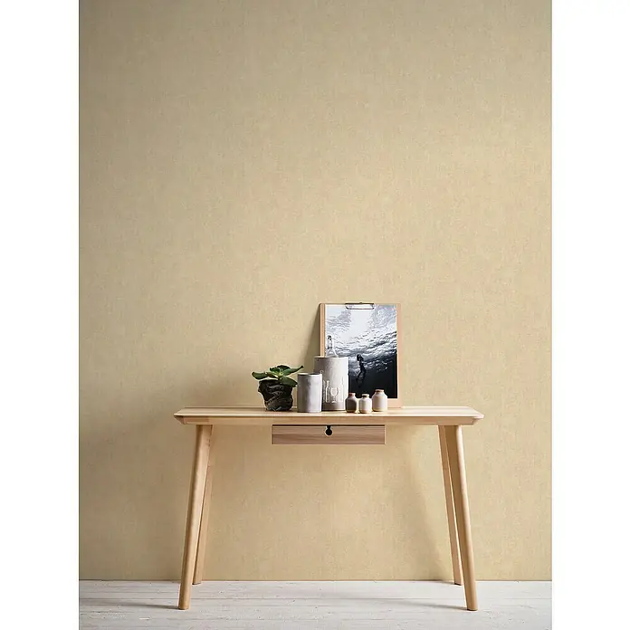 A.S. Création Hygge 2 Vliestapete - Beige, Uni, 10,05 x 0,53 m - Ein heller Holztisch mit Schublade, dekoriert mit Pflanzen, einer Flasche und einem gerahmten Bild, vor einem beigen Hintergrund.