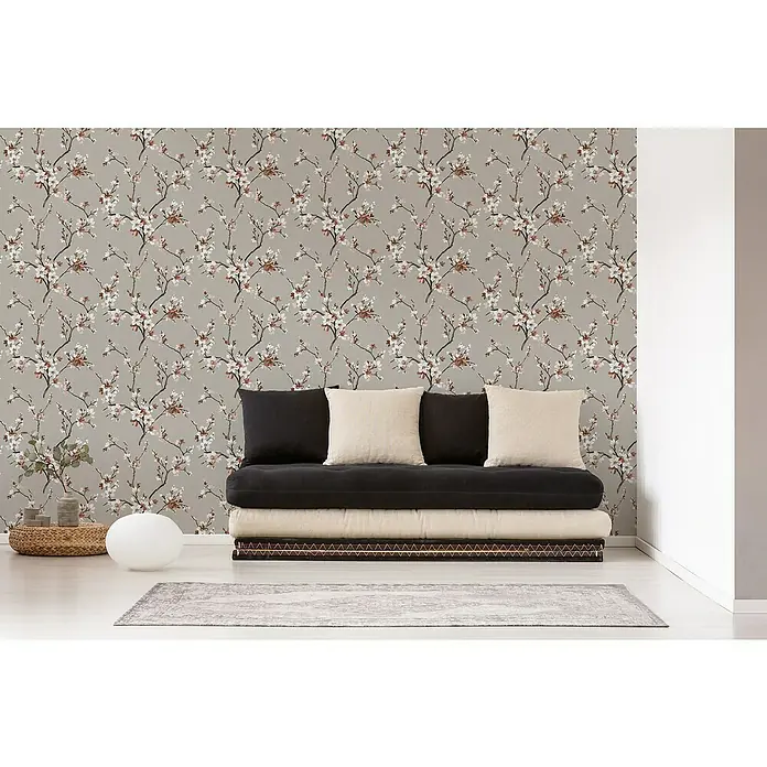 A.S. Création Desert Lodge Vliestapete Blütenranken - Grau/Rosè, Floral, 10,05 x 0,53 m - Ein gemütliches Zimmer mit einem stilvollen Futon-Sofa, das mit schwarzen und beigen Kissen dekoriert ist, vor einer grauen floralen Tapete.