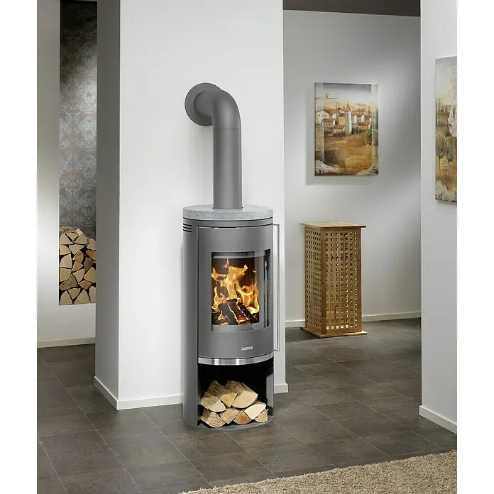 Justus Kaminofen Faro 2.0 - 6 kW, Raumheizvermögen: 115 m³, Verkleidung: Gusseisen/Speckstein, Speckstein - Moderner Justus Kaminofen mit Holzscheiten und Feuer. Der Kaminofen ist grau und steht auf einem grauen Fliesenboden.