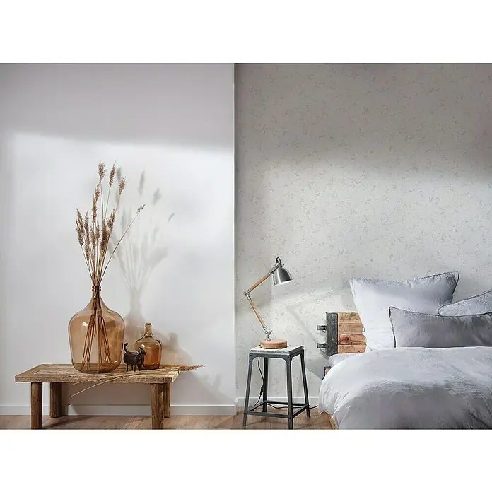 A.S. Création Attractive Vliestapete - Weiß/Grau, Uni, 10,05 x 0,53 m - Einrichtungsidee mit rustikalem Holztisch, Glasvasen, Dekorationen und einer Lampe auf einem Beistelltisch neben einem Bett.
