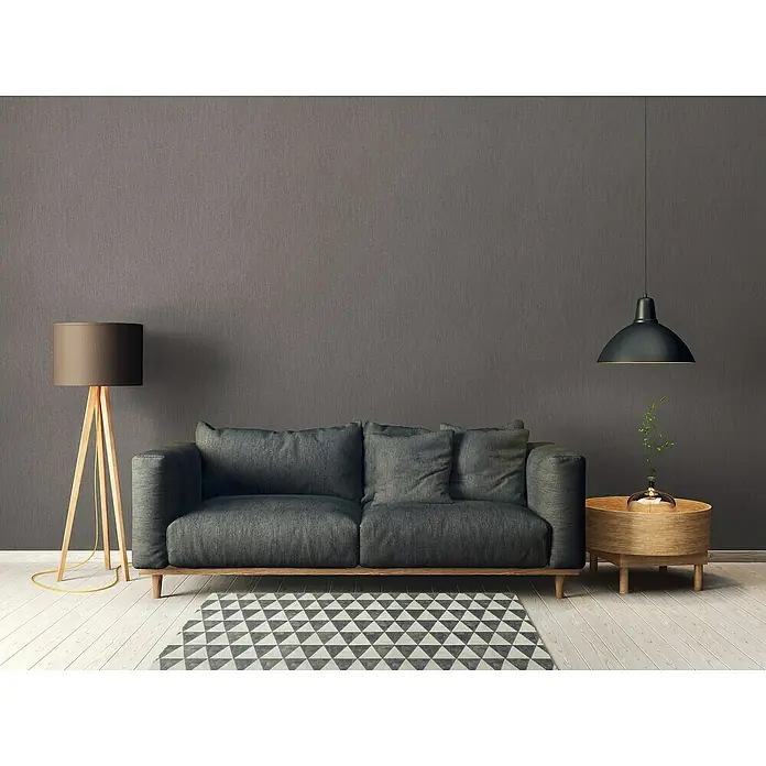 A.S. Création Daniel Hechter 6 Vliestapete Textilstruktur - Braunbeige, Uni, 10,05 x 0,53 m - Ein stilvolles graues Sofa mit passenden Kissen, einer Stehlampe und einem schwarzen Hängelampe über einem Holztisch.