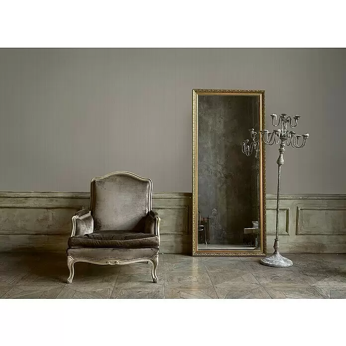 A.S. Création Mata Hari Vliestapete - Beige/Braun, Uni, 10,05 x 0,53 m - Ein brauner Samtsessel, ein hoher goldgerahmter Spiegel und ein großer silberner Kerzenleuchter in einem antiken Raum.