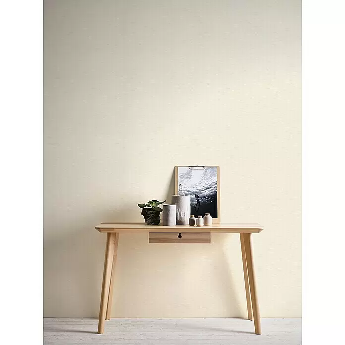 A.S. Création Hygge 2 Vliestapete Gewebe - Creme, Uni, 10,05 x 0,53 m - Texturierte, hellcremefarbene Wandverkleidung mit feinem Webmuster, die eine Wand hinter einem Holztisch schmückt.