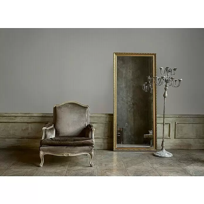 A.S. Création Mata Hari Vliestapete - Beige, Uni, 10,05 x 0,53 m - Ein Vintage-Sessel, ein verzierter Spiegel und ein hoher Kerzenständer in einem Raum mit Holzboden und Wandvertäfelung für eine klassische Ästhetik.