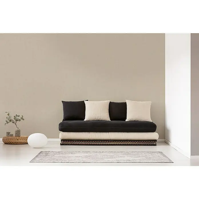 A.S. Création Titanium 3 Vliestapete - Pastelles Beige, Uni, 10,05 x 0,53 m - Bequemes Futon-Sofa mit schwarzen und cremefarbenen Kissen auf einem gemusterten Teppich. Minimalistisches Design für entspanntes Wohnen.