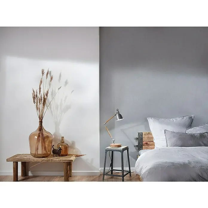 A.S. Création Titanium 3 Vliestapete - Wohnliches Grau, Uni, 10,05 x 0,53 m - Dekorative Glasvase mit Trockenblumen auf einem Holztisch, neben einer Lampe und einem Bett, die eine gemütliche und stilvolle Atmosphäre schaffen.