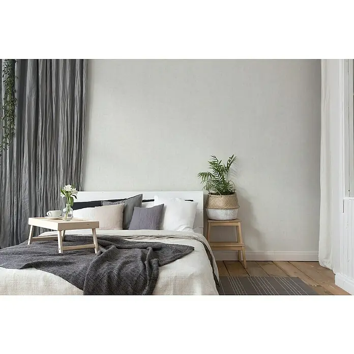 A.S. Création Titanium 3 Vliestapete - Strukturiertes Weiß, Uni, 10,05 x 0,53 m - Schlafzimmer mit einem Bett, das mit einer grauen Decke, Kissen und einem Nachttisch mit Pflanzen dekoriert ist. Vorhang und Tisch auf dem Bett.