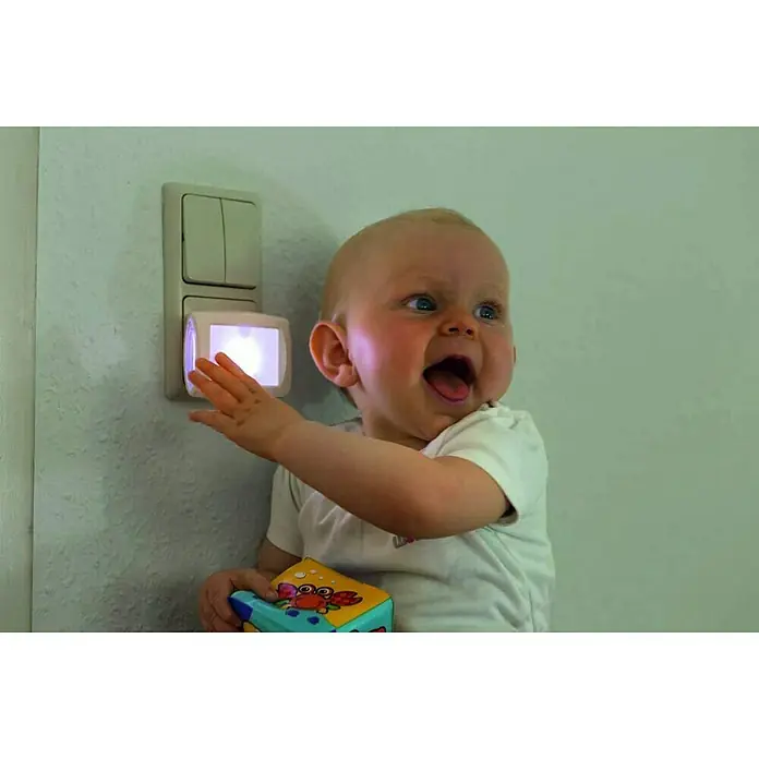 Abus Nachtlicht Lori - Weiß - Ein Baby das nach einem beleuchteten Nachtlicht an der Wand greift, es hält einen Spielzeugwürfel in der Hand.