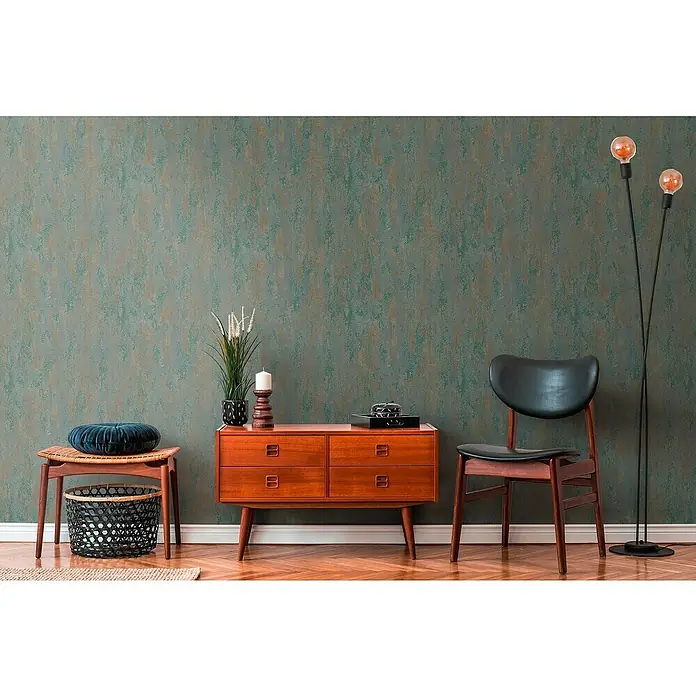 A.S. Création Trendwall 2 Vliestapete Rost - Rostiges Türkis, Metalloptik, 10,05 x 0,53 m - Ein Interieur mit einem Teakholz-Sideboard, einem Stuhl und einer Stehlampe vor einer strukturierten Wand. Dekorative Elemente ergänzen die Szene.