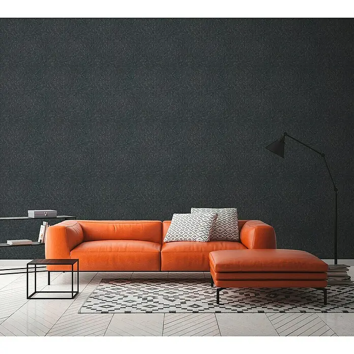 A.S. Création Titanium 3 Vliestapete Dynamischer 3D-EffektSchwarz, 3D-Optik, 10,05 x 0,53 m - Orangefarbenes Ledersofa mit passendem Ottoman, akzentuiert durch dekorative Kissen und eine Stehlampe, vor einer dunklen, gemusterten Wand.