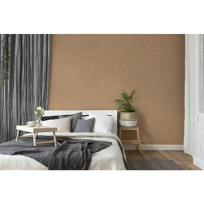 A.S. Création Desert Lodge Vliestapete Gewebestruktur - Ocker, Uni, 10,05 x 0,53 m - Ein gemütliches Schlafzimmer mit einem Bett, das mit einer grauen Decke und Kissen dekoriert ist, vor einer braunen Wand mit Textur.