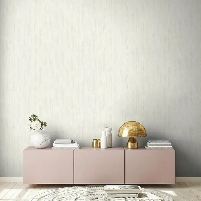 A.S. Création Hygge 2 Vliestapete Bäume - Creme/Beige, Floral, 10,05 x 0,53 m - Einrichtung mit einer zartrosa Kommode, dekoriert mit einer goldenen Lampe, Vasen und Büchern vor einer strukturierten Wand.