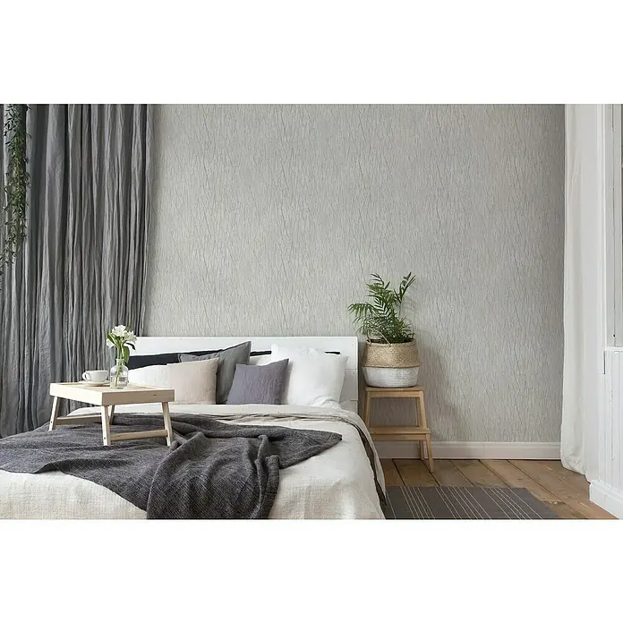 A.S. Création Hygge 2 Vliestapete Crush - Grau/Silber, Grafisch, 10,05 x 0,53 m - Helles Schlafzimmer mit einem Bett, das mit Kissen und einer Decke dekoriert ist, und einem stilvollen Nachttisch mit Pflanzen.