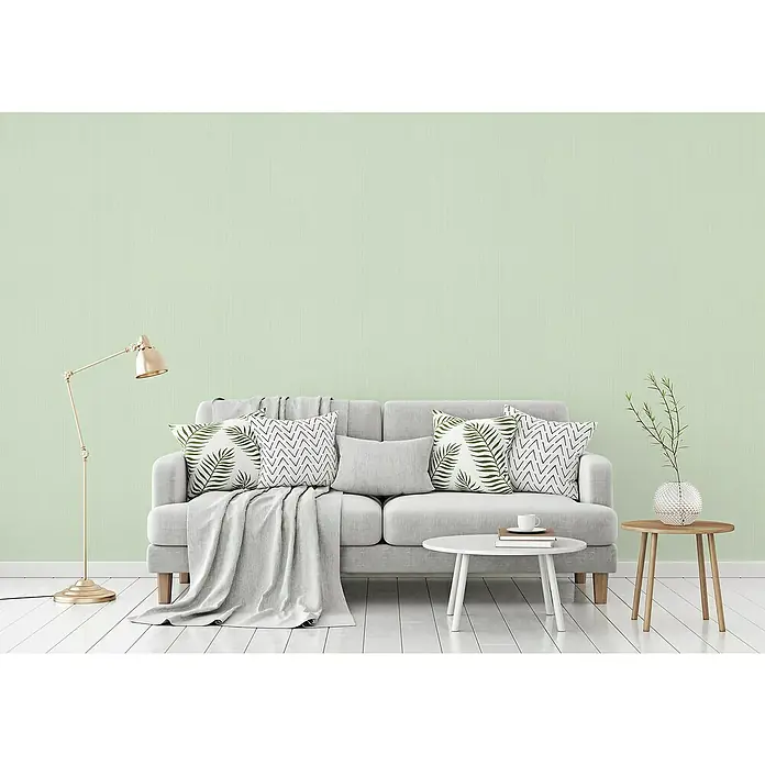 A.S. Création Michalsky 4 Vliestapete - Hellgrün, Uni, 10,05 x 0,53 m - Wohnzimmer-Szene mit grauem Sofa, Kissen mit Blattmuster, goldener Lampe und weißen Tischen, die eine gemütliche und moderne Atmosphäre ausstrahlen.