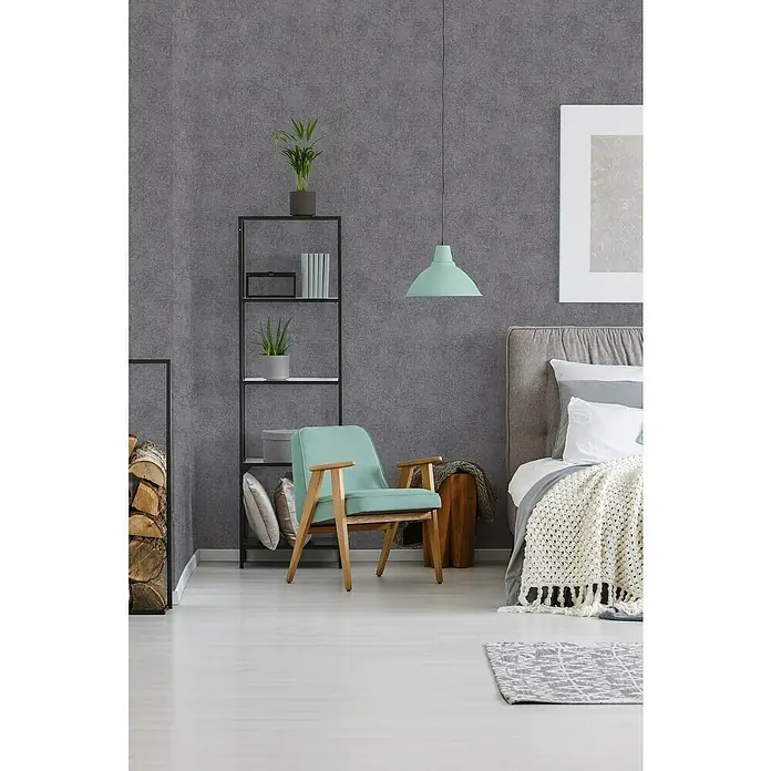A.S. Création Trendwall 2 Vliestapete - Grau/Bronze, Uni, 10,05 x 0,53 m - Einrichtungsidee mit hellblauem Sessel aus Holz, Bücherregal, Bett mit Decke und grauem Teppich auf hellem Boden vor grauer Wand.