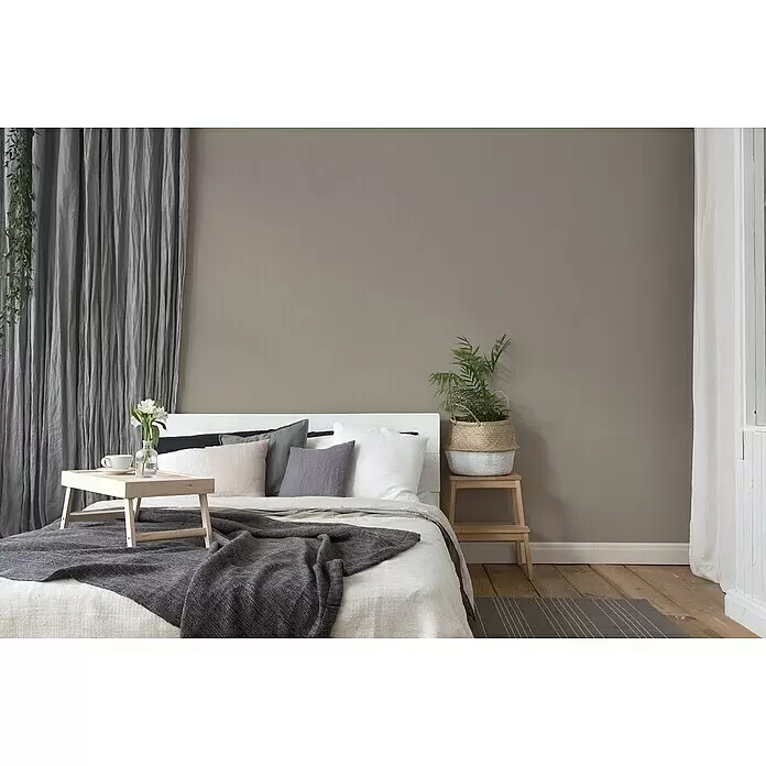 A.S. Création Desert Lodge Vliestapete - Braun, Uni, 10,05 x 0,53 m - Ein Bett mit einem weißen Bettbezug, verschiedenen Kissen in neutralen Tönen und einer dunkelgrauen Strickdecke.