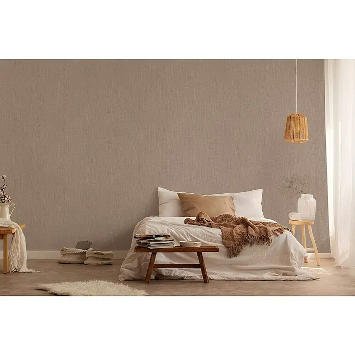 A.S. Création Daniel Hechter 6 Vliestapete Textilstruktur - Braun, Uni, 10,05 x 0,53 m - Schlafzimmer-Szene mit einem Bett mit beige-farbiger Tagesdecke, einem Holzhocker mit Büchern und einer dekorativen Hängelampe über dem Nachttisch.