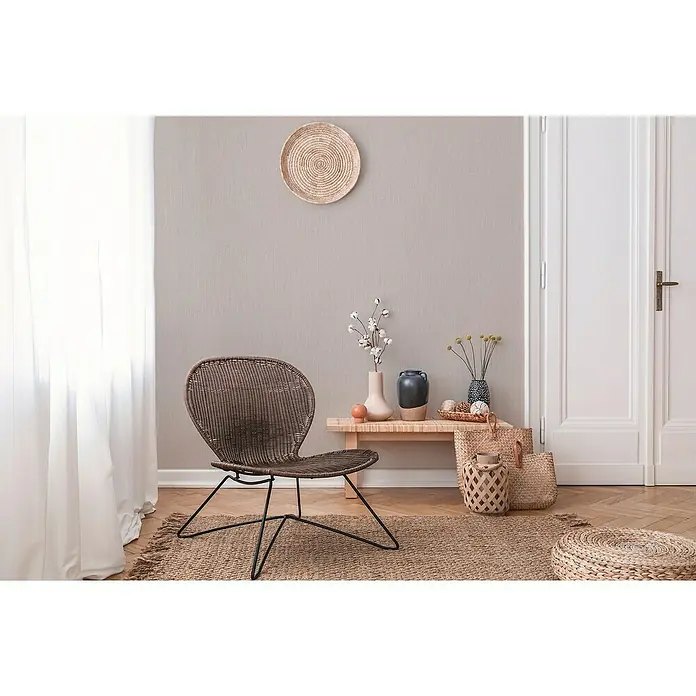 A.S. Création Daniel Hechter 6 Vliestapete Textilstruktur - Hellbeige, Uni, 10,05 x 0,53 m - Einrichtungsidee mit einem braunen Rattansessel auf einem Teppich, neben einem Holztisch mit Deko und zwei Körben.