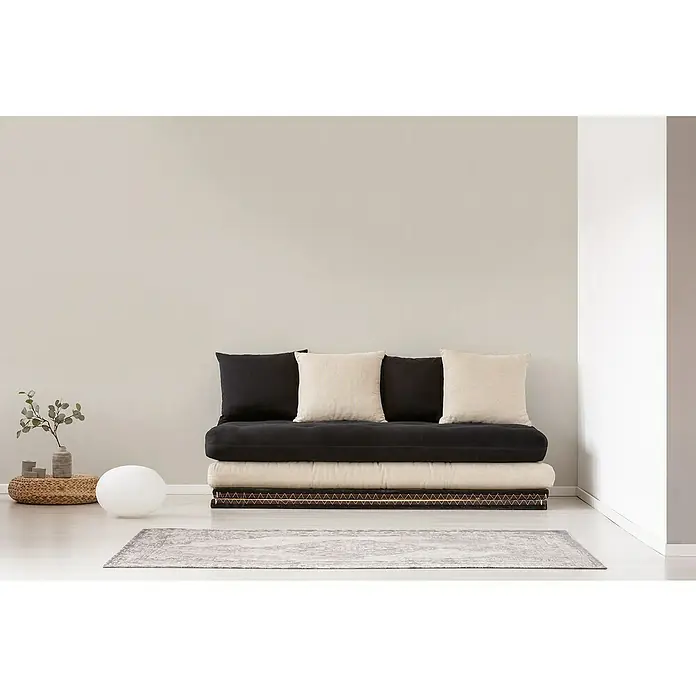 A.S. Création Desert Lodge Vliestapete - Kieselgrau hell, Uni, 10,05 x 0,53 m - Ein stilvolles Futonsofa mit schwarzen und beigen Kissen, platziert auf einem gemusterten Teppich in einem hellen Raum.