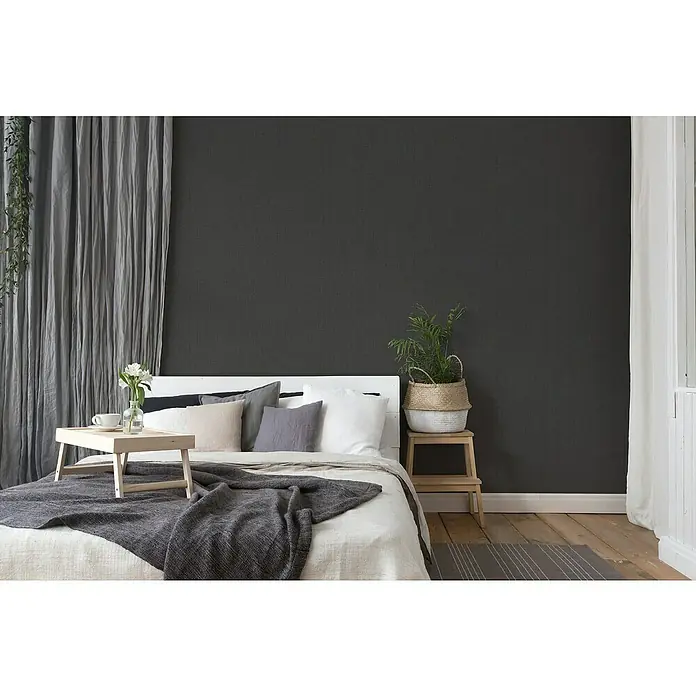 A.S. Création Desert Lodge Vliestapete - Schwarz, Uni, 10,05 x 0,53 m - Schlafzimmer mit Bett, Nachttisch, Pflanze, Vorhängen und Holztablett. Das graue Farbschema sorgt für eine ruhige Atmosphäre.