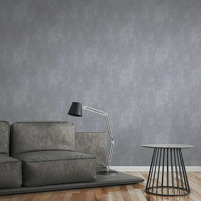 A.S. Création Titanium 3 Vliestapete I - Dunkelgrau, Betonoptik, 10,05 x 0,53 m - Eine moderne Wohnzimmerszene mit einem grauen Sofa, einer Stehlampe und einem Beistelltisch, die eine entspannte Atmosphäre schaffen.