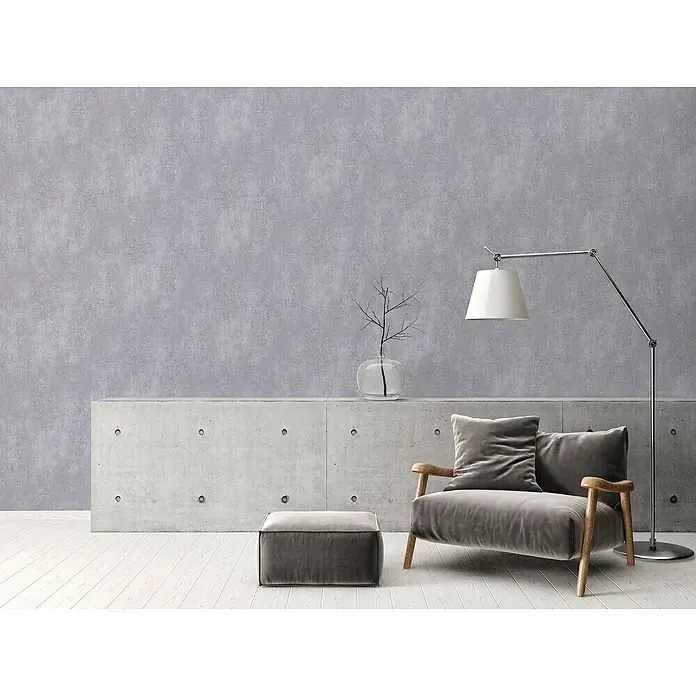 A.S. Création Titanium 3 Vliestapete I - Dunkelgrau, Betonoptik, 10,05 x 0,53 m - Elegantes Wohnzimmer mit einem grauen Samt-Sessel, einem passenden Hocker und einer modernen Stehlampe. Die schlichte Einrichtung wird durch eine minimalistische Dekoration ergänzt.