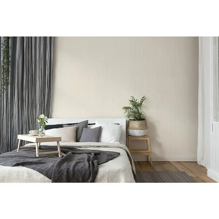 A.S. Création Hygge 2 Vliestapete Bäume - Creme/Beige, Floral, 10,05 x 0,53 m - Schlafzimmer mit großem Bett, das mit vielen Kissen und einer grauen Decke geschmückt ist. Ein kleiner Tisch steht auf dem Bett mit Blumen.