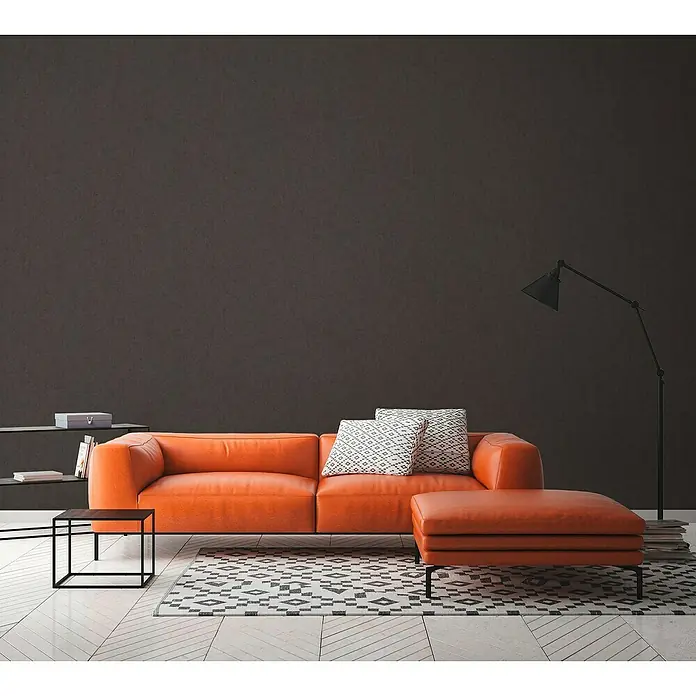 A.S. Création Titanium 3 Vliestapete - Dunkles Schwarz, Uni, 10,05 x 0,53 m - Ein modernes Wohnzimmer mit einem orangefarbenen Ledersofa, Kissen und einem Hocker auf einem gemusterten Teppich.