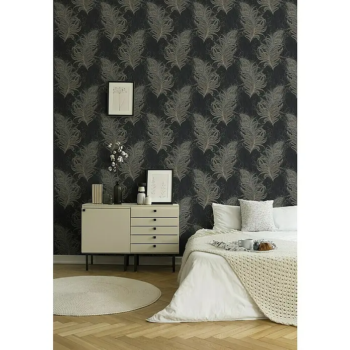 A.S. Création Mata Hari Vliestapete Federn - Schwarz/Grau, Tieroptik, 10,05 x 0,53 m - Schlafzimmer-Interieur mit einem stilvollen Nachttisch, einer runden Matte und einem Bett mit weißer Bettwäsche vor einer gemusterten Tapete.