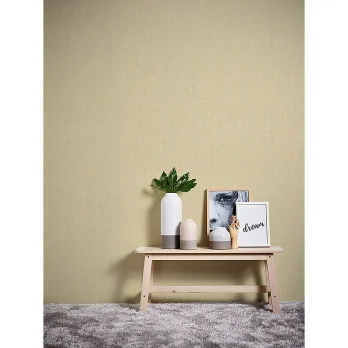 A.S. Création Hygge 2 Vliestapete Gewebe - Beige, Uni, 10,05 x 0,53 m - Beige Wandtapete mit einer kleinen Holzbank, auf der Vasen, ein Bild und eine Holzhand stehen. Weicher, grauer Teppich.