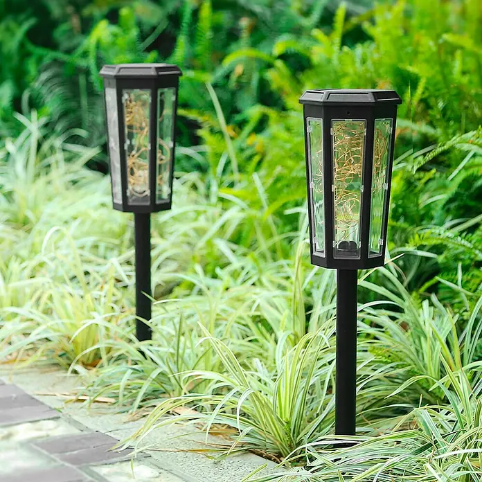 Arte confort Lámpara decorativa solar Aura - Negro, IP44 - Dos luces solares con bombillas están en el jardín rodeadas de vegetación, agregando un acento acogedor y energéticamente eficiente.