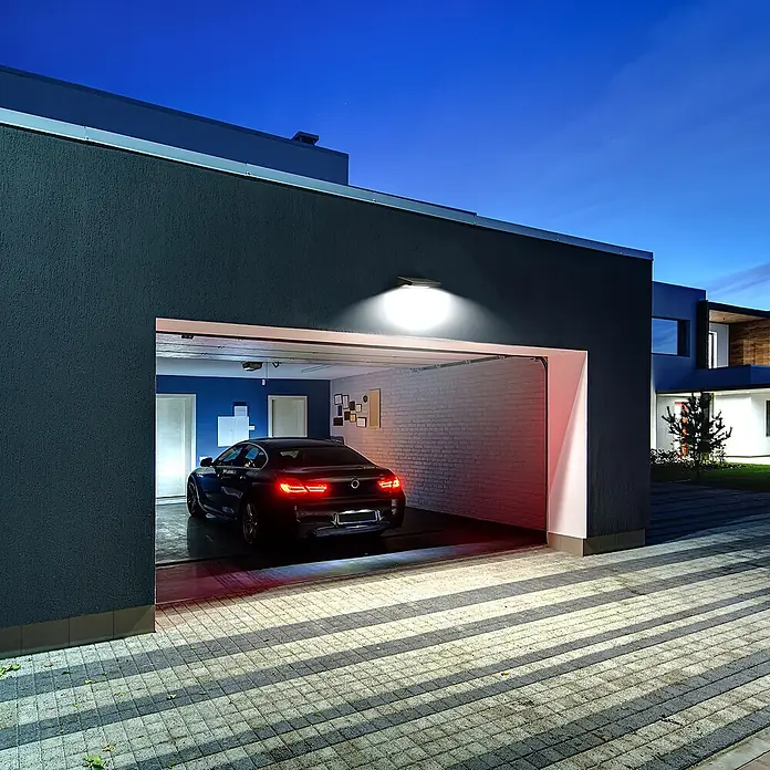 Arte confort Aplique solar para exterior Fenix - 9 W, L x An x Al: 16 x 22 x 7,9 cm, Gris, IP54, 1.300 lm - Vista exterior de un garaje con una luz LED solar que ilumina la entrada, con un coche negro en el interior.