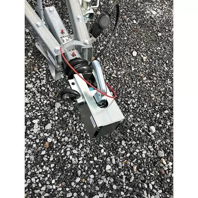 Basi Diebstahlsicherung KFZ 114 - Aluminium, Verzinkt - Silberne Anhängerkupplung mit schwarzem Gummibalg und rotem Sicherungskabel, auf Kies liegend.