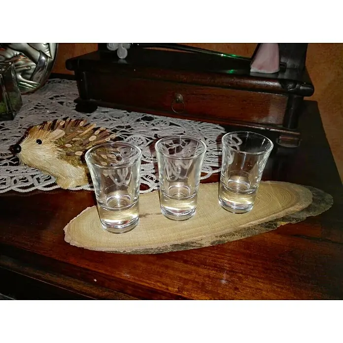 Disco de tronco de madera Oval - Ø x Es: 15 cm x 10 mm - Tres vasos de chupito vacíos sobre una tabla de madera rústica, presentados de forma decorativa en una mesa de madera oscura.