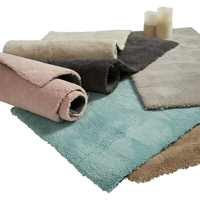 Alfombra de pelo corto Toscana - Gris, 300 x 200 cm, 100% Poliéster - Esta imagen muestra una selección de alfombras de baño suaves en varios colores, enrolladas y extendidas, junto con velas decorativas.